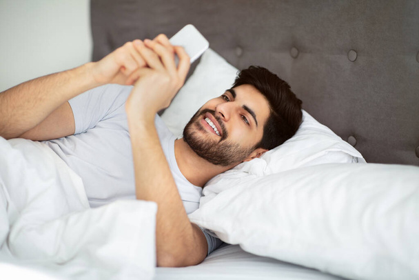 Social networks, morning good news, positive message at weekend. Cheerful arab man waking up and looking at smartphone - Foto, Bild