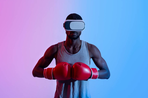 Ισχυρός μαύρος αθλητής σε VR ακουστικά βάζοντας γάντια πυγμαχίας μαζί σε νέον φως - Φωτογραφία, εικόνα