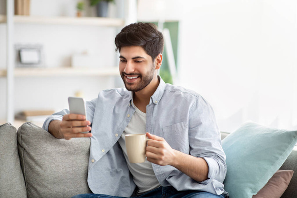 Online life concept. Positive arab man holding modern smartphone and cup of coffee at home, free space - Foto, afbeelding