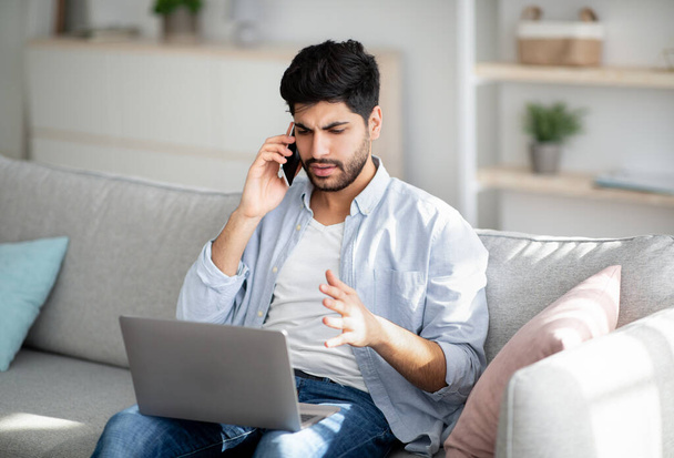 Problemas informáticos. Homem árabe descontente falando no smartphone e olhando na tela do laptop em casa - Foto, Imagem