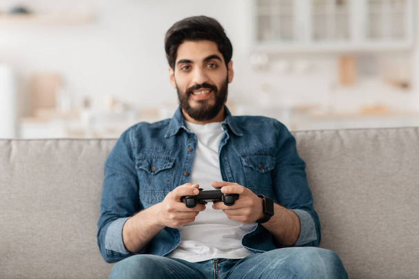 Расслабленный ближневосточный парень играет в видеоигры с джойстиком дома, сидит на диване, копирует пространство - Фото, изображение