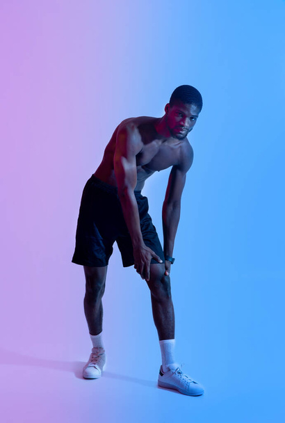 Voller Länge junger schwarzer Sportler mit nacktem Oberkörper, der sein verletztes Knie berührt und unter Schmerzen in Neonbeleuchtung leidet - Foto, Bild