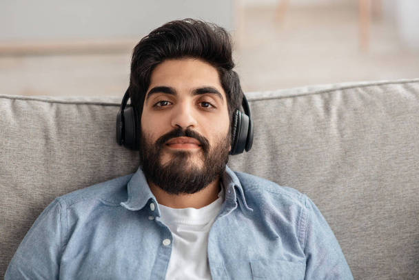 Closeup portrait of bearded arab guy in wireless headset reclining on sofa and looking at camera, copy space - Φωτογραφία, εικόνα