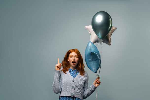 Ginger excite woman pointing finger upward while posing with balloons isolated over grey background - Fotoğraf, Görsel