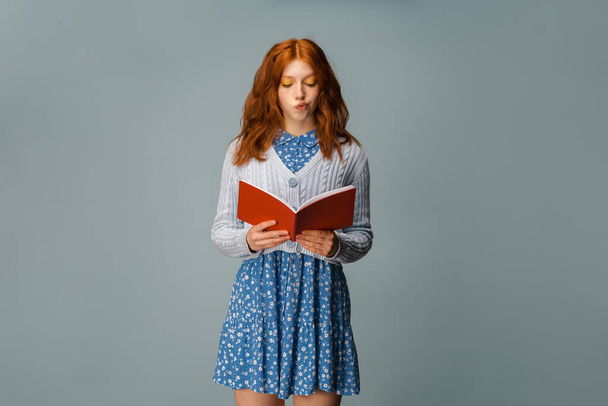 Young ginger puzzled woman posing while reading her diary isolated over grey background - 写真・画像