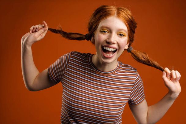 Ginger joyful woman making fun with her tails and laughing isolated over orange background - Фото, изображение