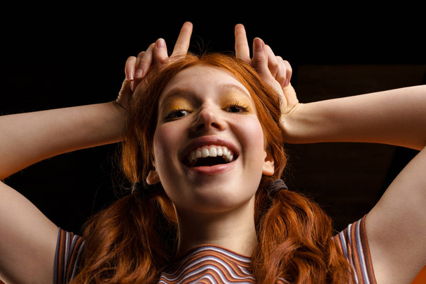 Young ginger amusing woman smiling and showing horns with fingers isolated over black background - Zdjęcie, obraz