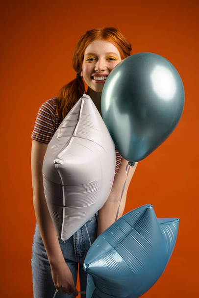 Ginger joyful woman smiling while posing with balloons isolated over orange background - Foto, Imagen