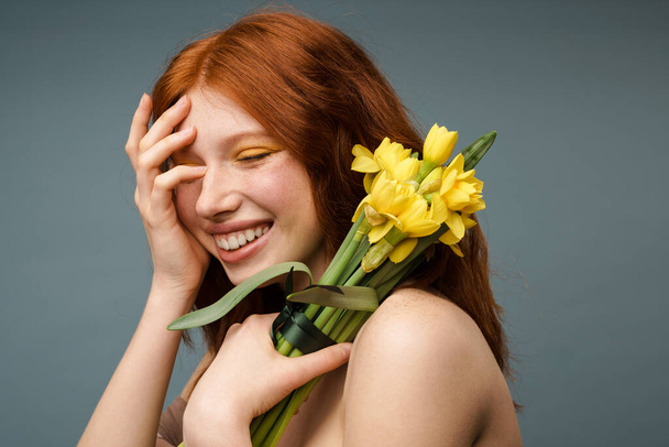 Shirtless ginger smiling woman posing with yellow flowers isolated over grey background - Φωτογραφία, εικόνα