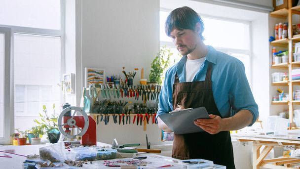Um jovem mestre conduzindo inventário em um estúdio criativo. Um homem de avental com prancheta - Foto, Imagem