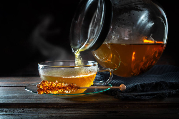 Hot steaming tea is poured from a pot into a glass cup, served with a stick of rock candy, motion blur, dark rustic wooden background with copy space, selected focus, narrow depth of field - Photo, image