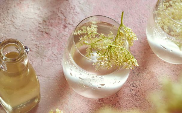 A glass of lemonade and homemade herbal syrup with fresh elder flowers on bright pink background - Фото, изображение