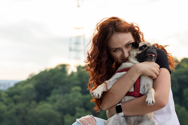 Giovane rossa donna abbraccia il suo cane carlino nel parco. - Foto, immagini