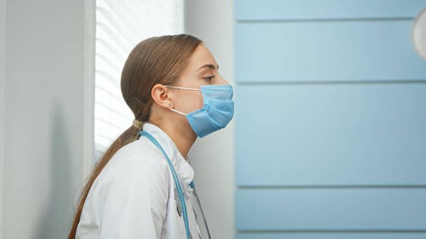 Erschöpfte junge Frau in weißem Mantel mit Stethoskop am Hals zieht blauen Einweg-Mundschutz ab - Foto, Bild