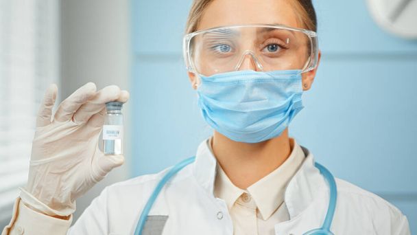 Молодая женщина-исследователь в синих одноразовых очках маски и белом халате держит прозрачный флакон с вакциной - Фото, изображение