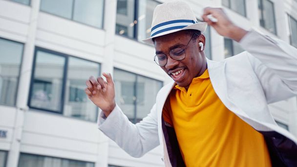 Afro-Amerikaanse man met hoed en draadloze koptelefoon luistert naar muziek en dansen met een vrolijke glimlach op megalopolis straat - Foto, afbeelding