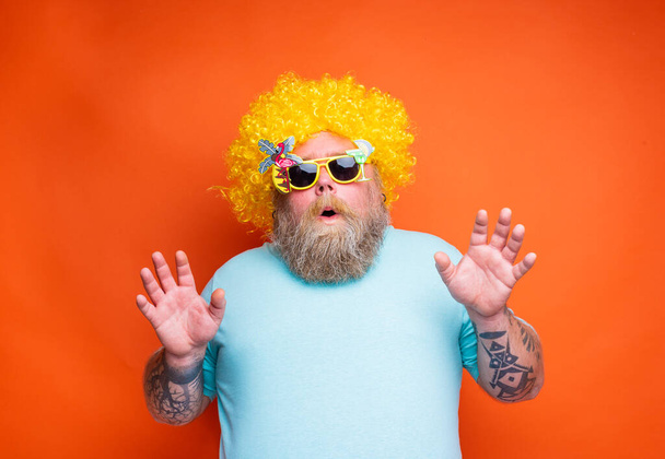 El hombre gordo asombrado con barba, tatuajes y gafas de sol se divierte con la peluca amarilla - Foto, imagen
