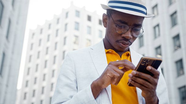 Hombre negro en traje con tipos de sombrero en el teléfono inteligente y ríe alegremente de pie junto a los edificios de oficinas - Foto, Imagen