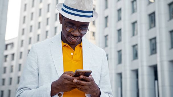 Sonriente hombre afroamericano en gafas y sombreros en el teléfono inteligente y sonríe con felicidad - Foto, Imagen