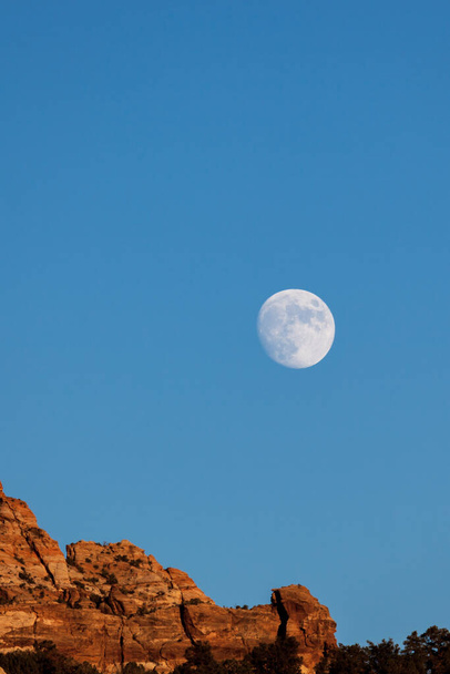 Una luna casi llena que se eleva por encima de las montañas de arenisca roja con un cielo azul por la noche cerca del Parque Nacional Zion, Utah. - Foto, imagen