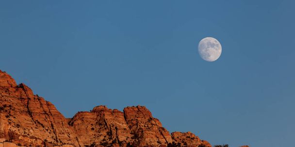 Una luna casi llena que se eleva por encima de las montañas de arenisca roja con un cielo azul por la noche cerca del Parque Nacional Zion, Utah. - Foto, imagen