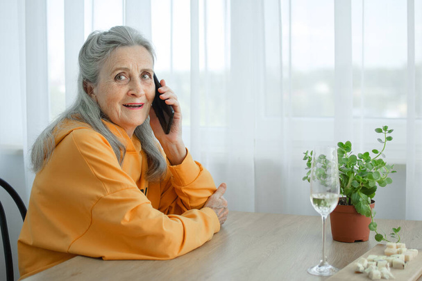 Красивая старая бабушка с седыми волосами и лицо с морщинами с помощью смартфона, разговаривая с кем-то и сидя за столом дома на фоне окна, День матери, счастливый выход на пенсию - Фото, изображение