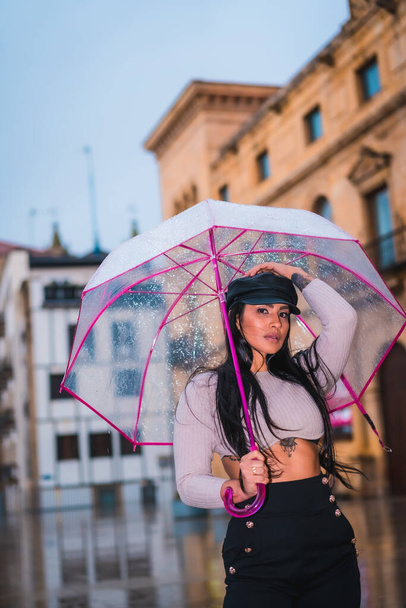 Posing of a young brunette Latina with a leather cap in the autumn rain with a transparent umbrella - Valokuva, kuva