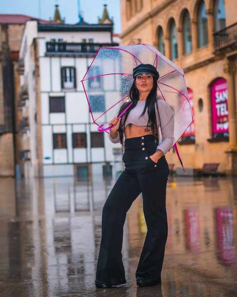Posing of a young brunette Latina with a leather hat in the rain with a transparent umbrella in the city - 写真・画像