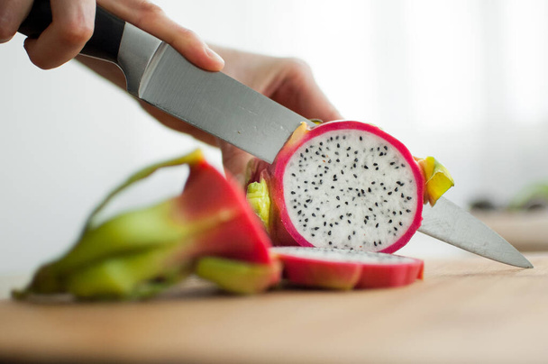 Female hands is cutting a dragon fruit or pitaya with pink skin and white pulp with black seeds on wooden cut board on the table. Exotic fruits, healthy eating concept - Foto, afbeelding