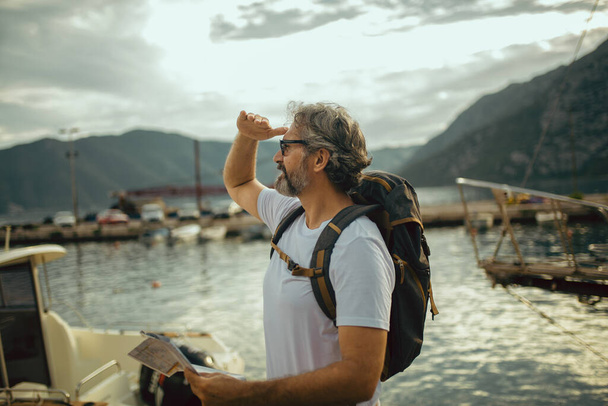 Sorrindo turista maduro homem de pé com mapa e mochila perto do mar. - Foto, Imagem