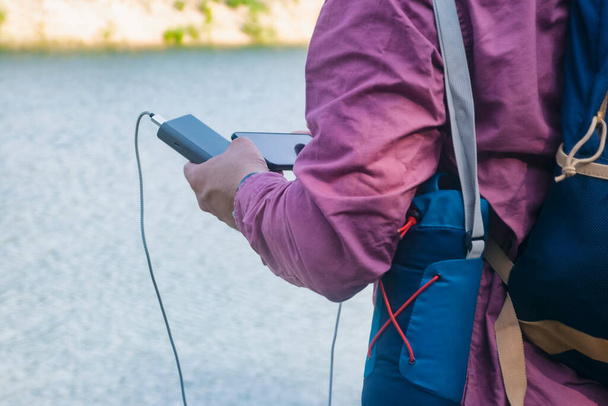 The guy is holding a portable charger with a smartphone in his hand. Man on a background of nature with a greenery and a lake. - Photo, Image