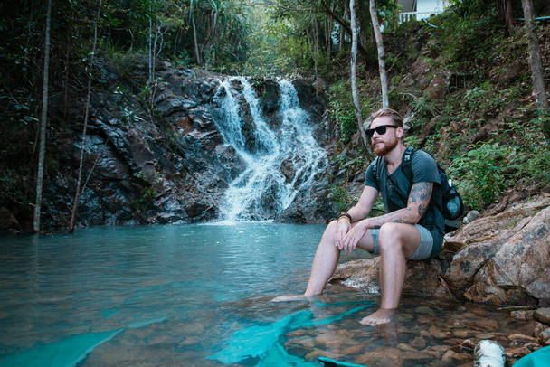 Male tourist near a small mountain tropical lake and waterfall. Young handsome caucasian guy at the foot of a waterfall on a hot day in a tropical forest - Photo, Image