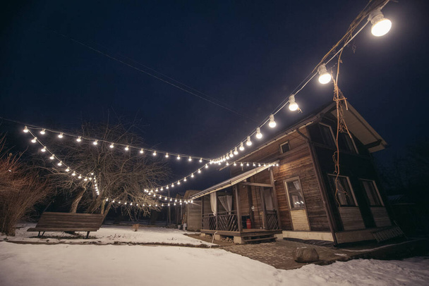 Casa de campo de madeira com uma varanda na parcela no inverno. Vista exterior. Iluminando o local com uma grinalda de lâmpadas. - Foto, Imagem