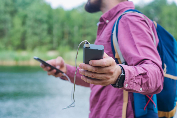 The guy is holding a portable charger with a smartphone in his hand. Man on a background of nature with a greenery and a lake. - Foto, Imagen