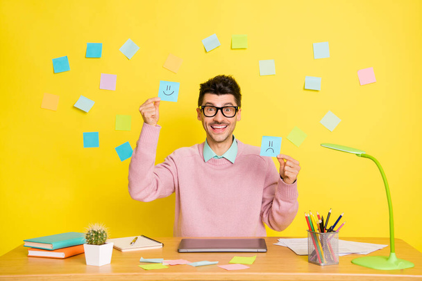 Photo of young cheerful smiling positive man in glasses showing happy and sad paper emoji isolated on yellow color background - Фото, зображення