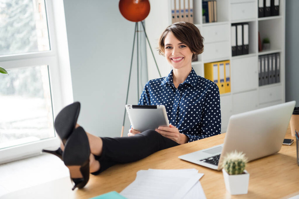 Portrait of attractive cheerful girl director sitting in chair putting legs on table using ebook at light workplace workstation indoors - Photo, image