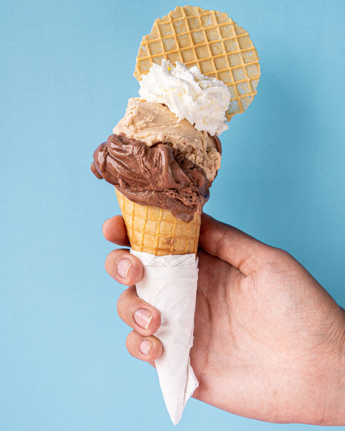 Cone de waffle requintado com gelato de chocolate, avelã e chantilly, acompanhado de um biscoito de waffle, segurado com uma mão, em um fundo azul. - Foto, Imagem