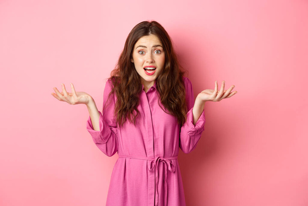 Confused young brunette woman with nervous face expression, raising hands up and shrugging shoulders, dont know, cant understand, standing over pink background - Photo, Image