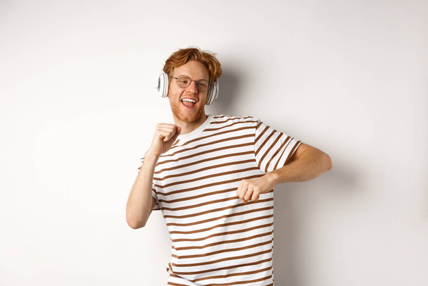 Technology concept. Happy redhead man listening music in headphones and dancing cheerful, standing in t-shirt against white background - Photo, Image
