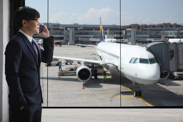 ビジネスマンは空港で携帯電話を呼んでる彼は立って飛行機を見てる  - 写真・画像