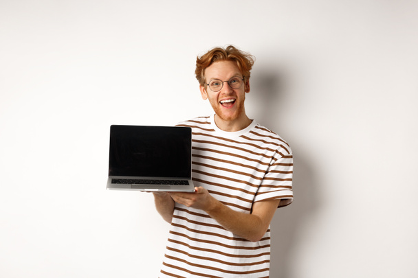 Αστείος κοκκινομάλλης με γυαλιά που δείχνει διαφήμιση οθόνης laptop και χαμογελά. Guy με κόκκινα μαλλιά επιδεικνύει promo ή banner στην οθόνη, λευκό φόντο - Φωτογραφία, εικόνα