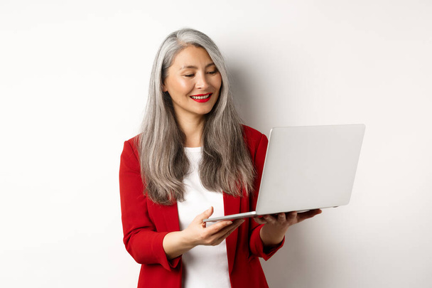 Interesy. odnosząca sukcesy azjatycka bizneswoman w czerwonej marynarce pracująca na laptopie, uśmiechnięta i czytająca ekran, stojąca na białym tle - Zdjęcie, obraz