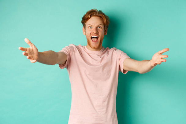 Jeune homme amical et heureux avec les cheveux roux, étirer les mains dans un accueil chaleureux, tendre les bras pour étreindre et sourire joyeusement, debout dans un t-shirt sur fond de menthe - Photo, image