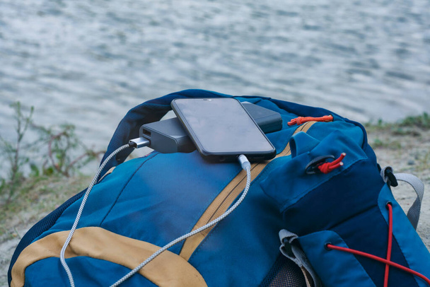 Smartphone is charged using a portable charger. Power Bank charges the phone outdoors with a backpack for tourism in the background of nature and the river - Foto, Imagem