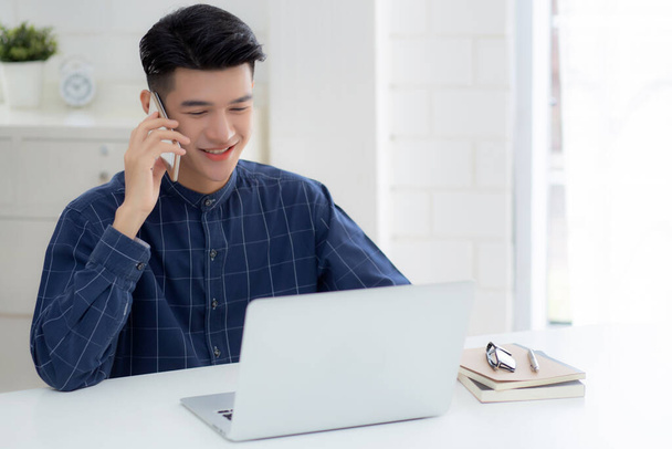 Mladý asijský muž mluví telefon a práci z domova s notebookem, na volné noze pomocí notebooku na internet on-line a mluvit na smartphone, domácí život, obchodní a komunikační koncept. - Fotografie, Obrázek