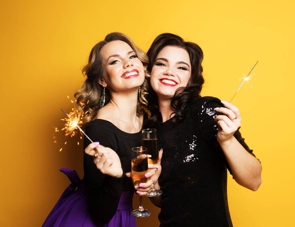 Portrait of two young women with a champagne glasses and sparklers over yellow background - Valokuva, kuva