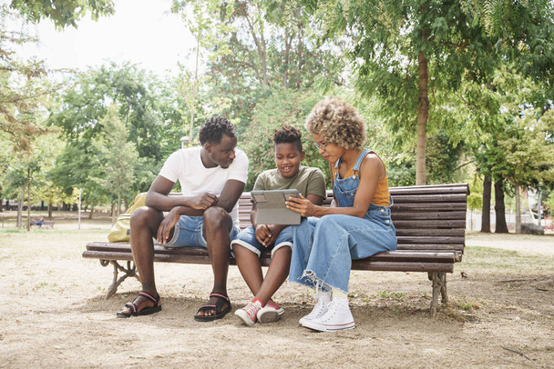 Familia afroamericana divirtiéndose con la tableta para ver videos en línea y redes sociales. jóvenes fresco millennial padres y lindo sonriente niño sentado en banco en al aire libre público parque - Foto, Imagen