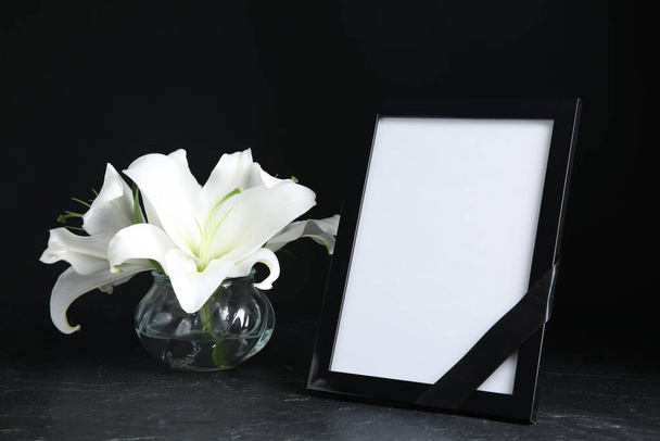 Funeral photo frame with ribbon and white lilies on black table against dark background. Space for design - Foto, imagen