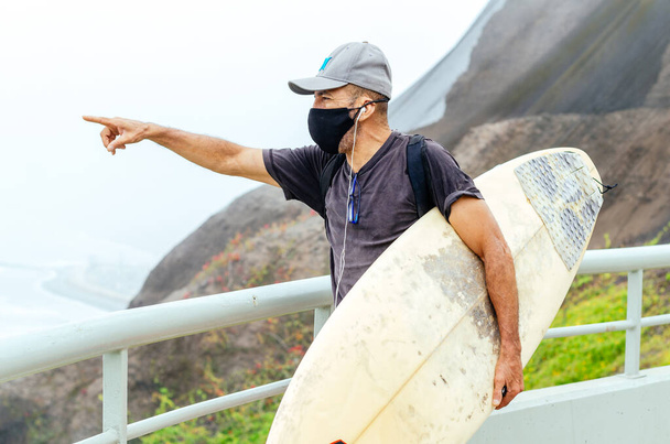Older surfer man with covid-19 protective mask and a surfboard - 写真・画像