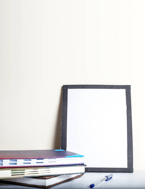 A pile of notebooks next to a pen and a blank picture with a black frame on a white background - Foto, Imagem